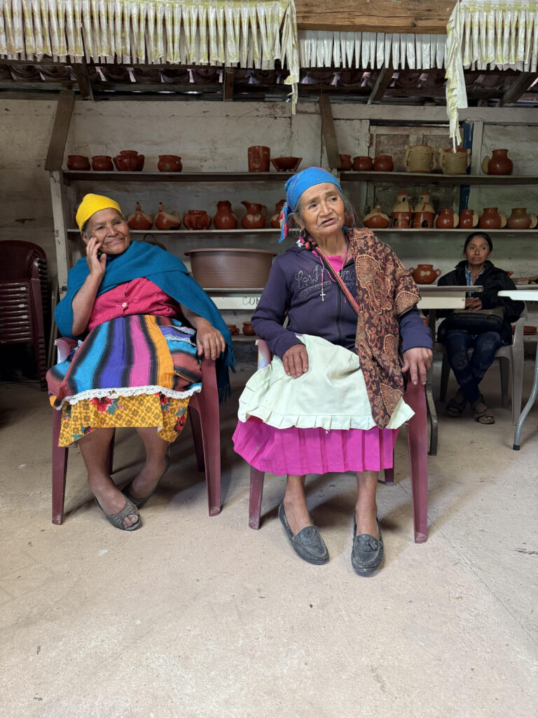 Two Indigenous women in brightly colored outfits sit on chairs in a room filled with pottery.