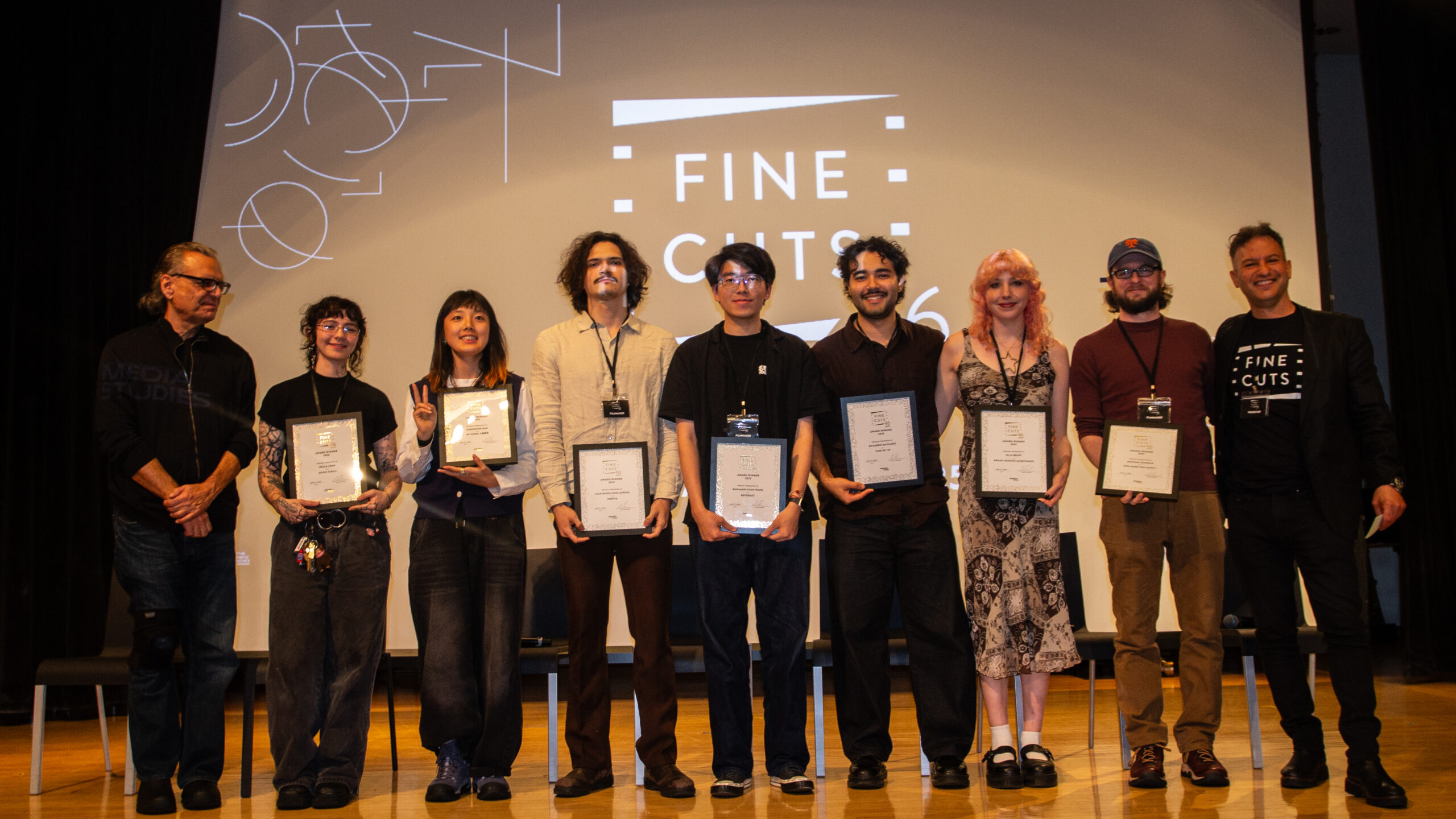 Nine people stand on stage, looking into the camera. The center seven people hold certificates in frames. The screen in the background reads “Fine Cuts.”
