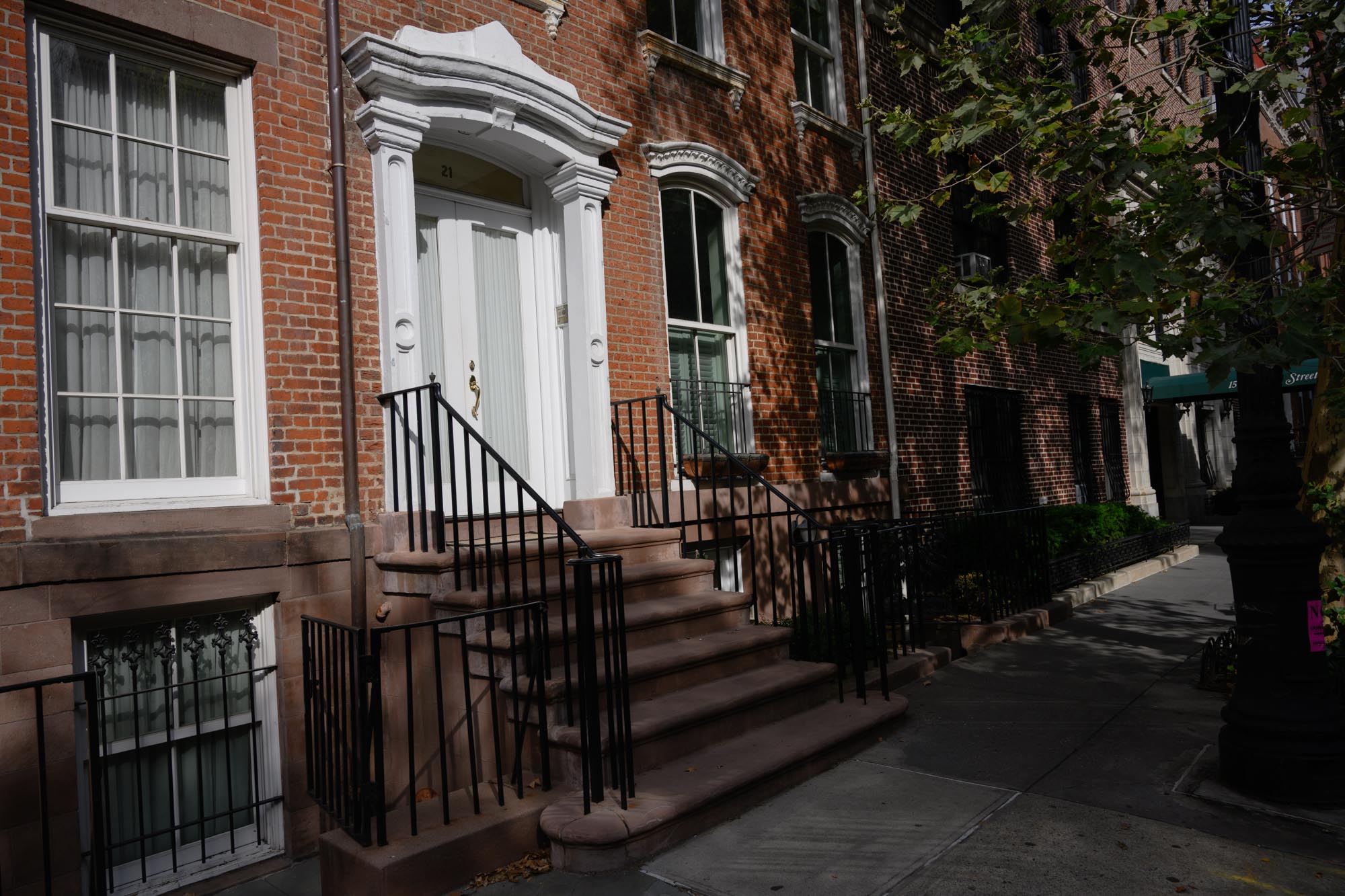 The photo shows a shaded exterior and the first floor of 21 W. 11th St., a brownstone with a stoop and wrought iron railings.