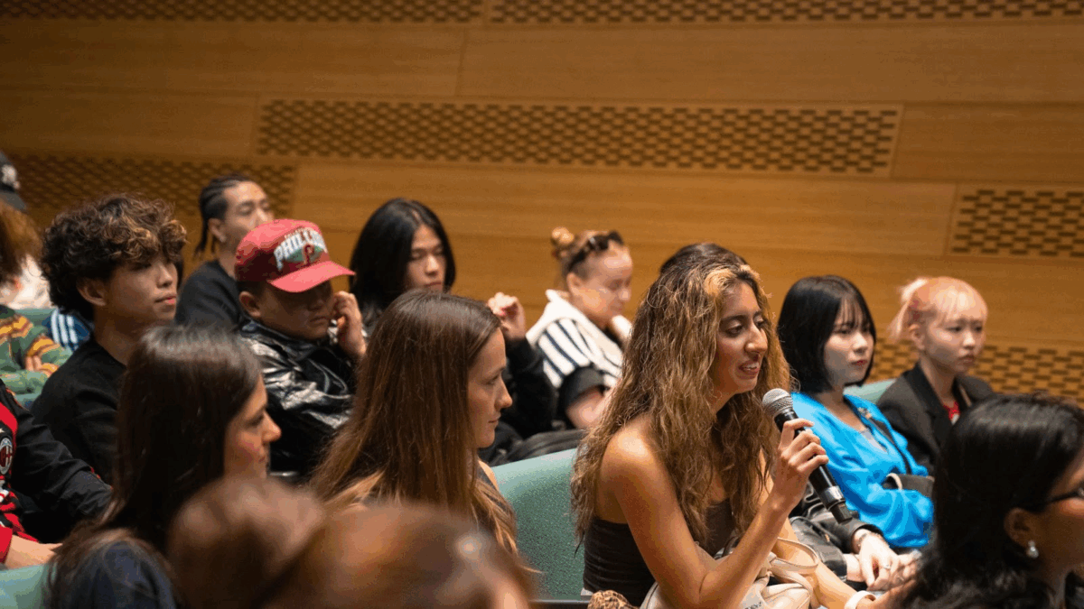  Audience with students in seats, looking forward to the panel. One student with brown hair holds a microphone, smiling. 