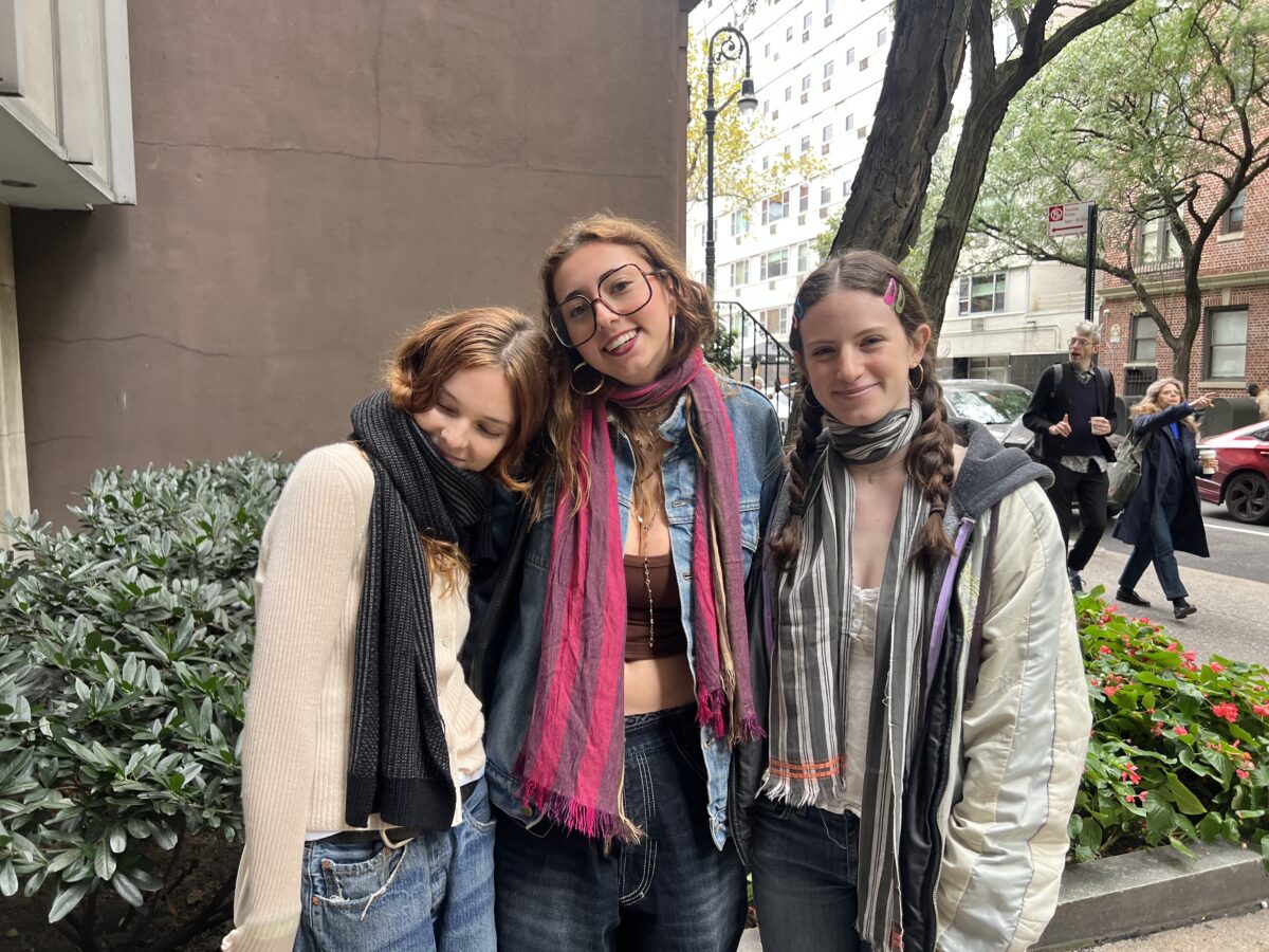  Three girls stand side by side. They’re all smiling, wearing different-colored, striped scarves. 

