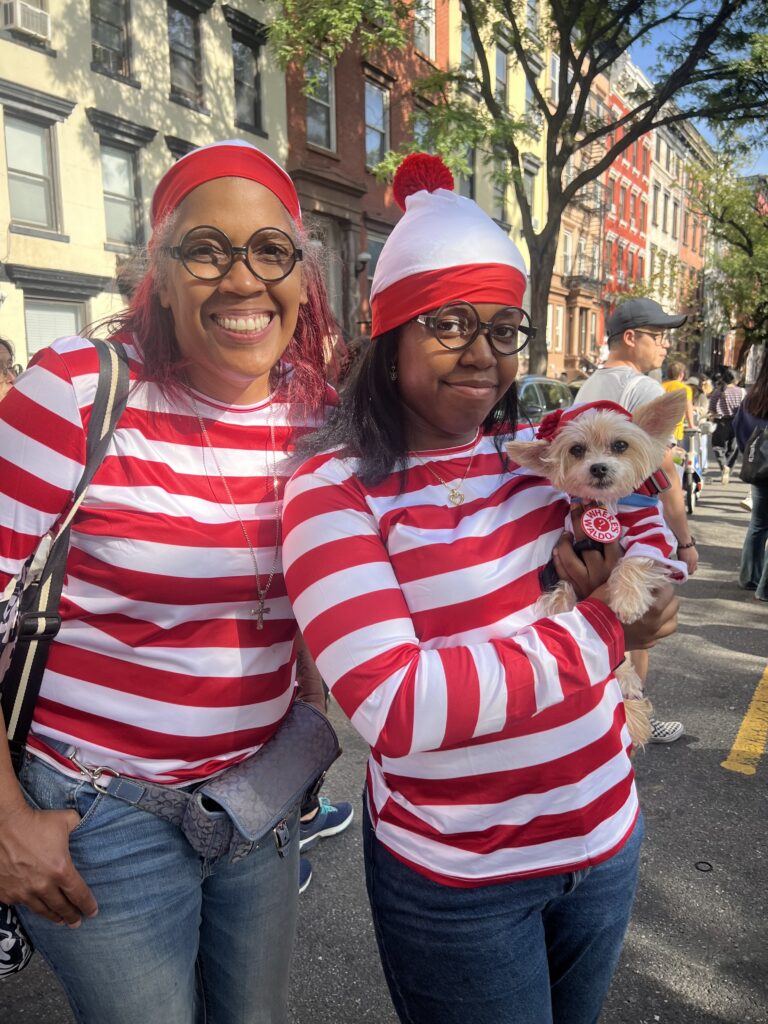 Two smiling people dressed in matching red-and-white striped “Where’s Waldo?” costumes, complete with round glasses and red hats, stand on a city street. One of them is holding a small dog also dressed in a red-and-white striped outfit.