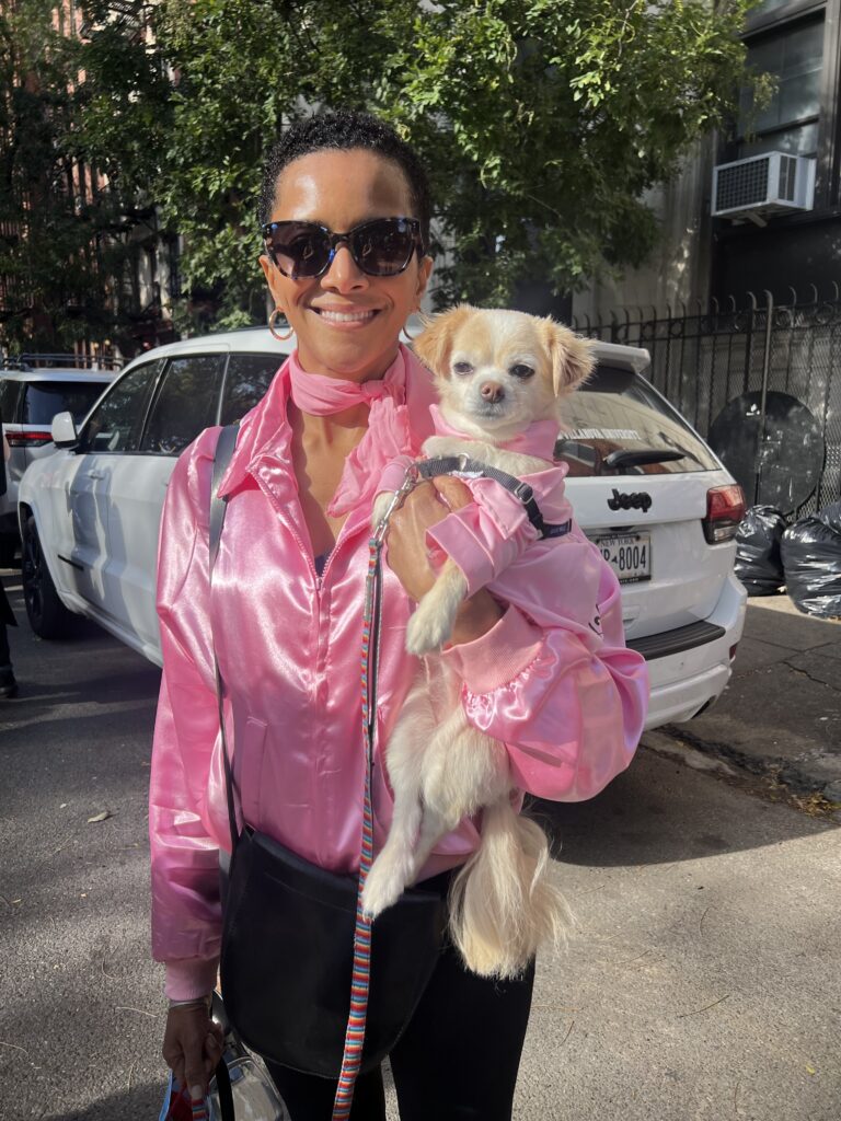 A smiling person wearing a shiny pink blouse, matching pink scarf, and sunglasses stands outdoors holding a small light-colored dog dressed in a pink outfit. They are standing on a sunny street with parked cars and trees in the background.