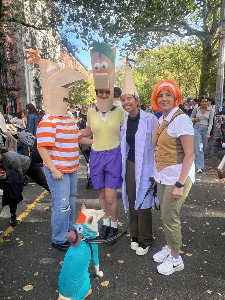 A group of four people dressed as characters from Phineas and Ferb pose together outdoors on a city street. Two wear large paper masks as Phineas and Ferb, one is dressed as Dr. Doofenshmirtz in a lab coat, and another as Candace with an orange wig. A dog in front of them is dressed as Perry the Platypus.