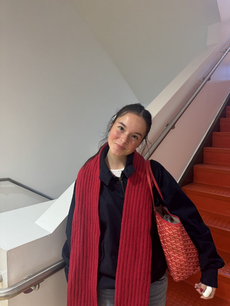 A girl stands in front of red stairs. She wears light denim jeans, a white t-shirt, a navy blue zip-up coat, and a bright red scarf. 
