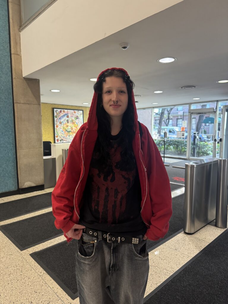  A student stands inside the lobby of Alvin Johnson/J.M. Kaplan Hall wearing a red zip-up hoodie over a black graphic tee, loose gray denim jeans, and a black studded belt.
