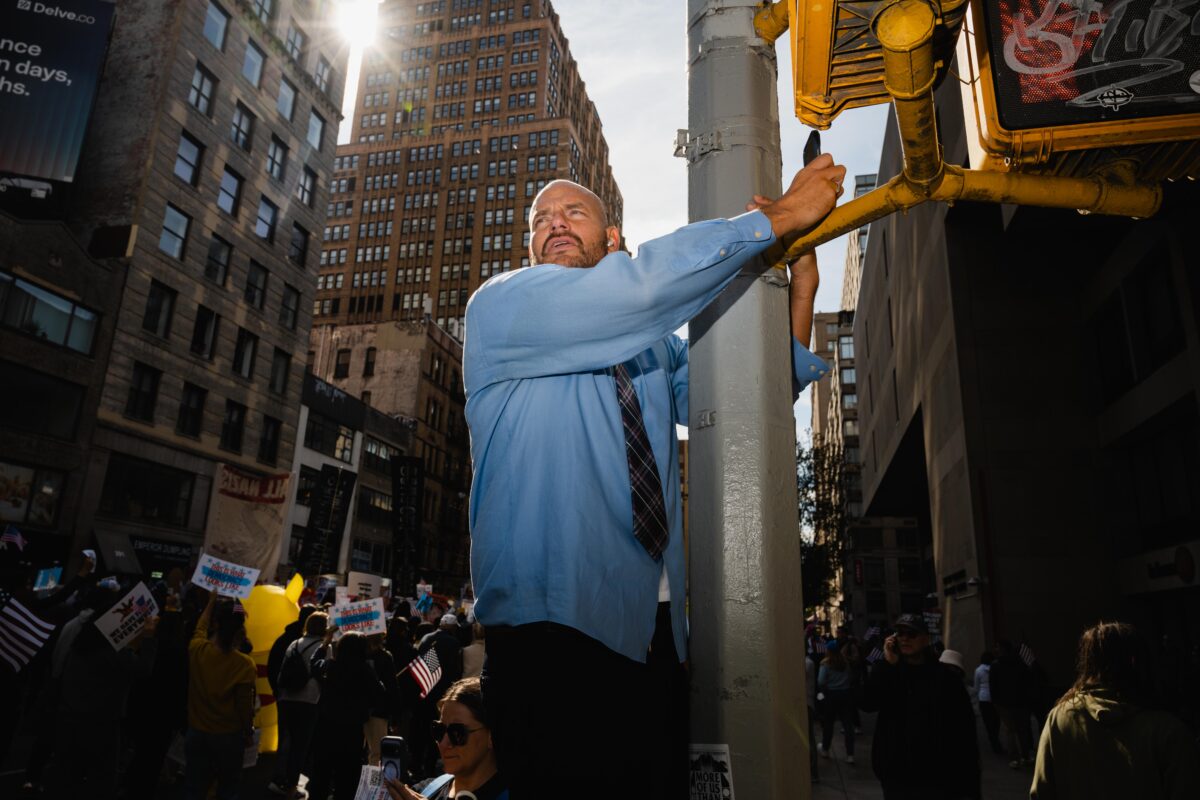 Man in blue shirt holding onto a street pole in an urban area with a protest behind him.