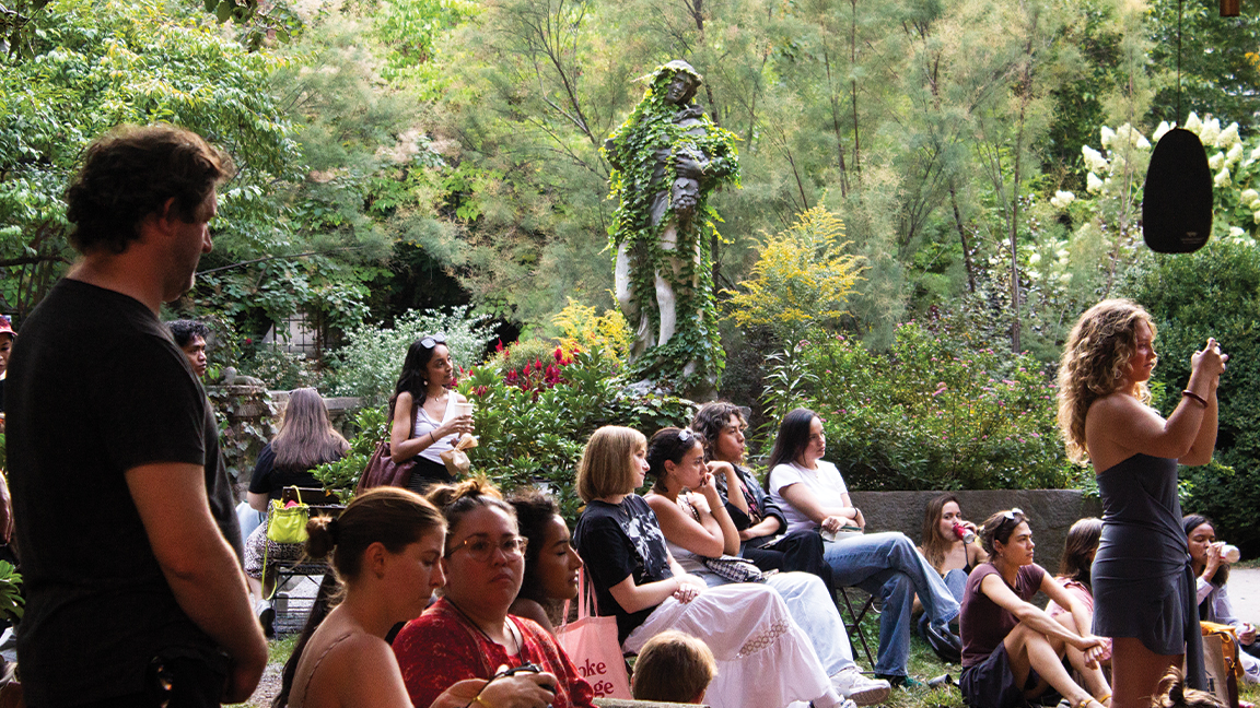 Community members sit outdoors in rows in front of a statue of a woman covered in vines.