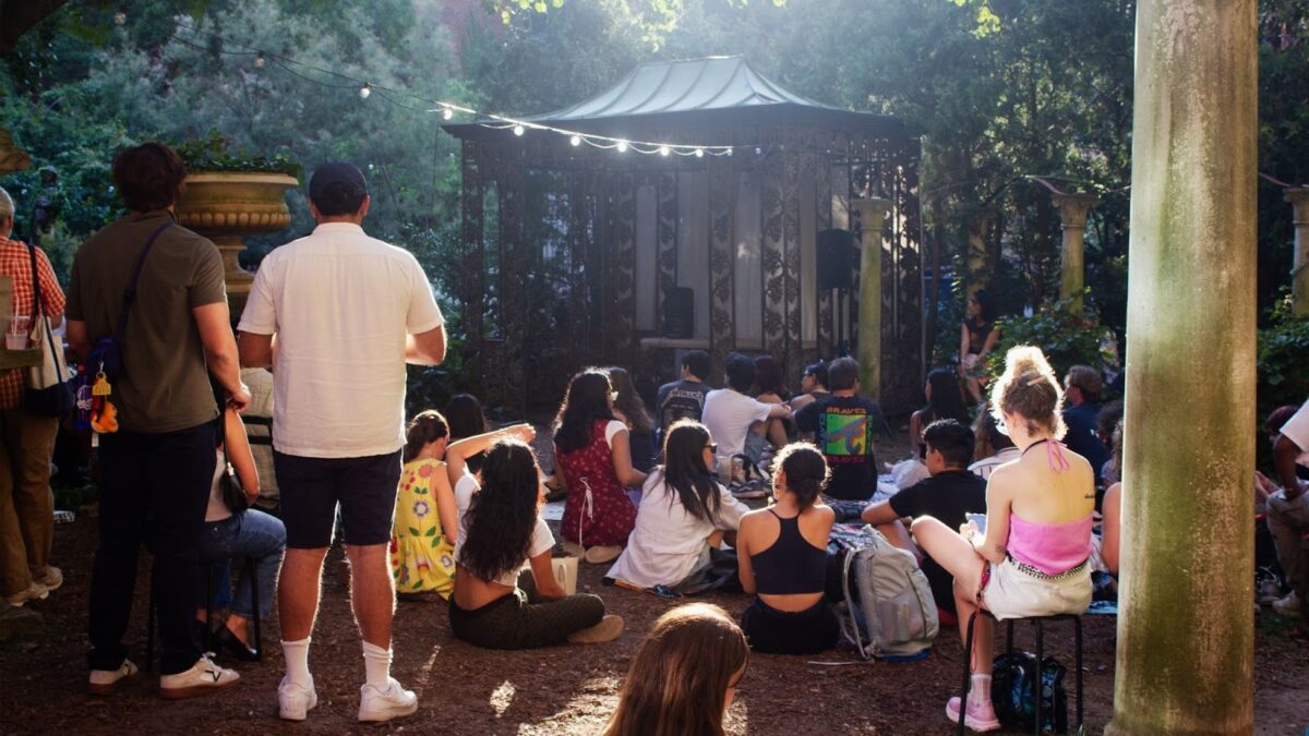 People sit in chairs and on the ground facing a small stage in the garden.