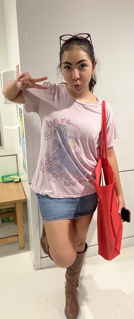 Full body picture of a girl wearing a pink graphic t-shirt; a blue denim skirt; knee-high, brown leather boots; glasses on top of her head; and a red bag over her left shoulder. 