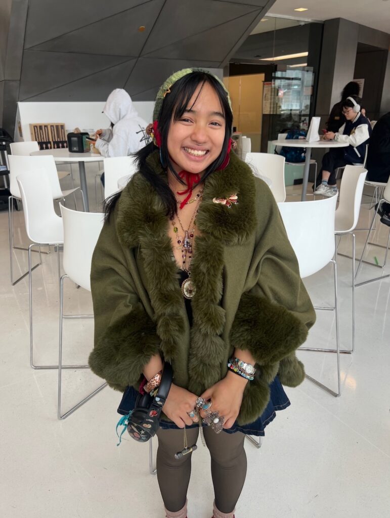 A smiling person in an olive-green fur-trimmed coat and knit bonnet stands indoors, wearing layered jewelry and holding decorated headphones, with people seated in the background.