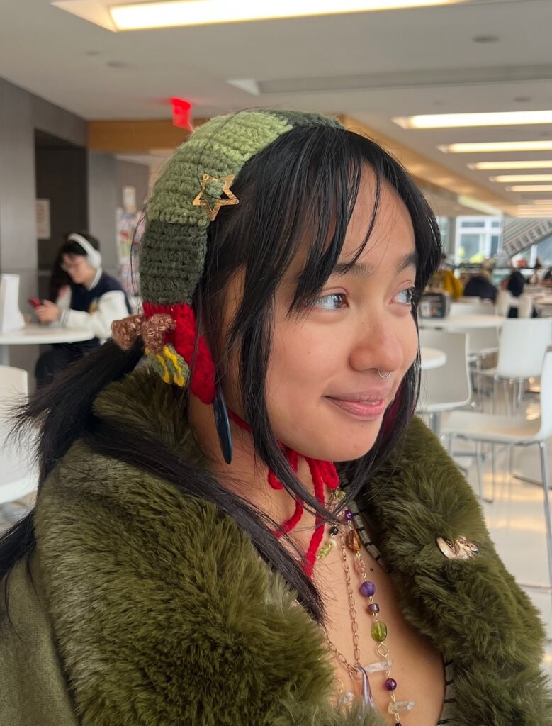 Close-up of a person in a green knit bonnet with a star pin and an olive faux-fur coat is shown in close-up, smiling slightly and wearing layered beaded necklaces in a bright indoor café.