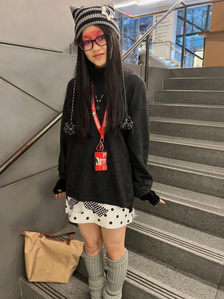 A person stands on an indoor staircase wearing a striped cat-ear beanie, glasses, a black oversized sweater, a white patterned skirt, gray leg warmers, and a red lanyard, with a tan bag placed on the step beside them.
