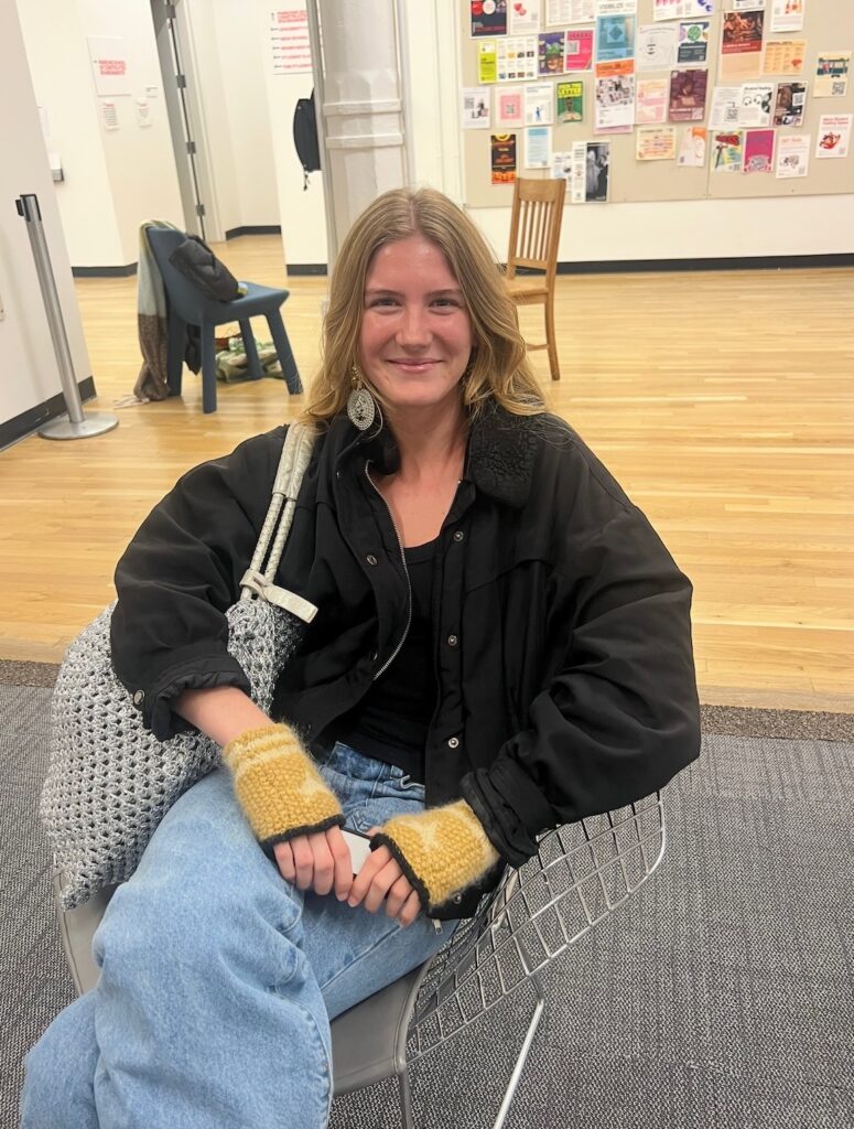 A person sits in a wire chair indoors, smiling and wearing a black jacket, light blue jeans, and yellow knit fingerless gloves, with a large crocheted bag over one shoulder and posters on the wall behind them.