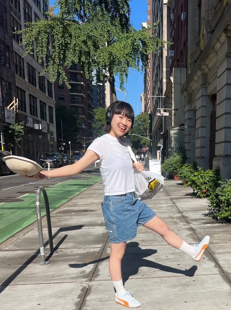 Girl posing on sidewalk wearing black bob, denim shorts, white socks in blue and orange sneakers, white top, and black headphones. She holds a white bag and food container.
