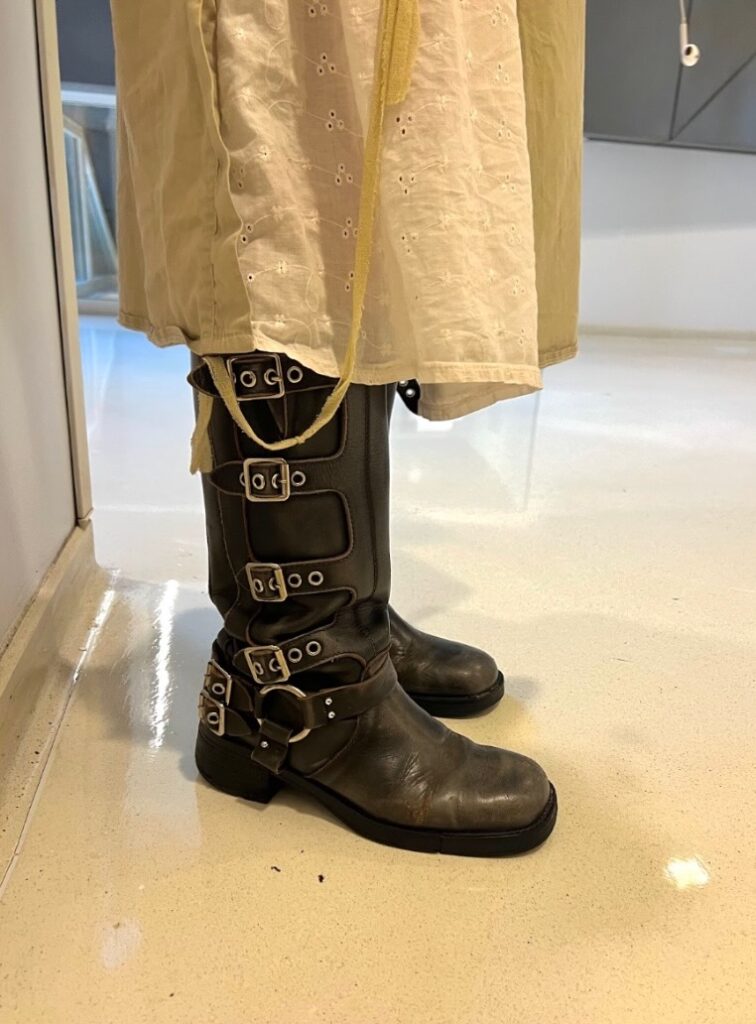 A close up of dark brown leather boots that go below the knee. There are five silver  buckles on the side of the boot. A tan and white skirt covers the top of the boots. 

