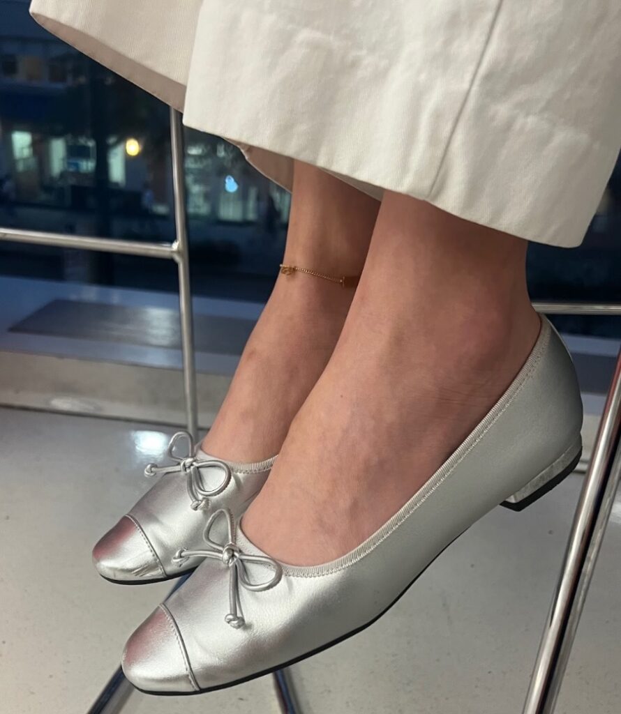 A close-up of silver ballerina flats with a very low heel. The bottom part of the leg is shown wearing white pants. The right ankle has a gold anklet.
