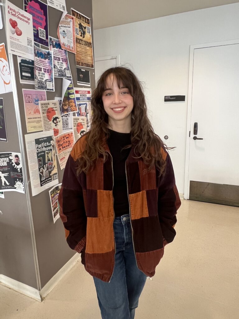A person stands indoors smiling, wearing a patchwork corduroy jacket in warm tones over a black shirt and jeans, with a wall covered in colorful posters behind them.
