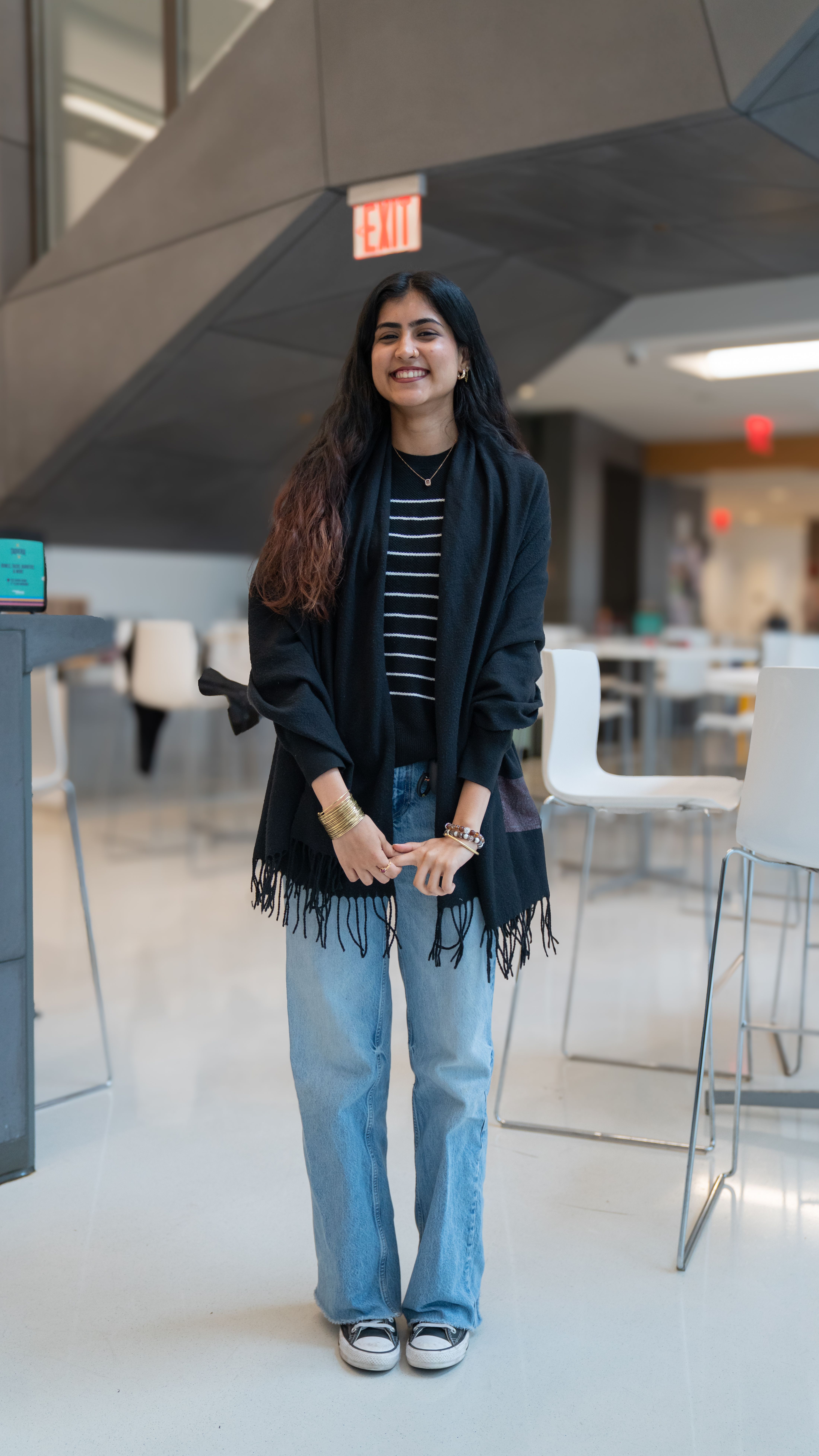 A girl standing indoors wearing a black shawl over a black and white striped top, light blue jeans, and black sneakers, smiling warmly in a modern space with white chairs and tables in the background.