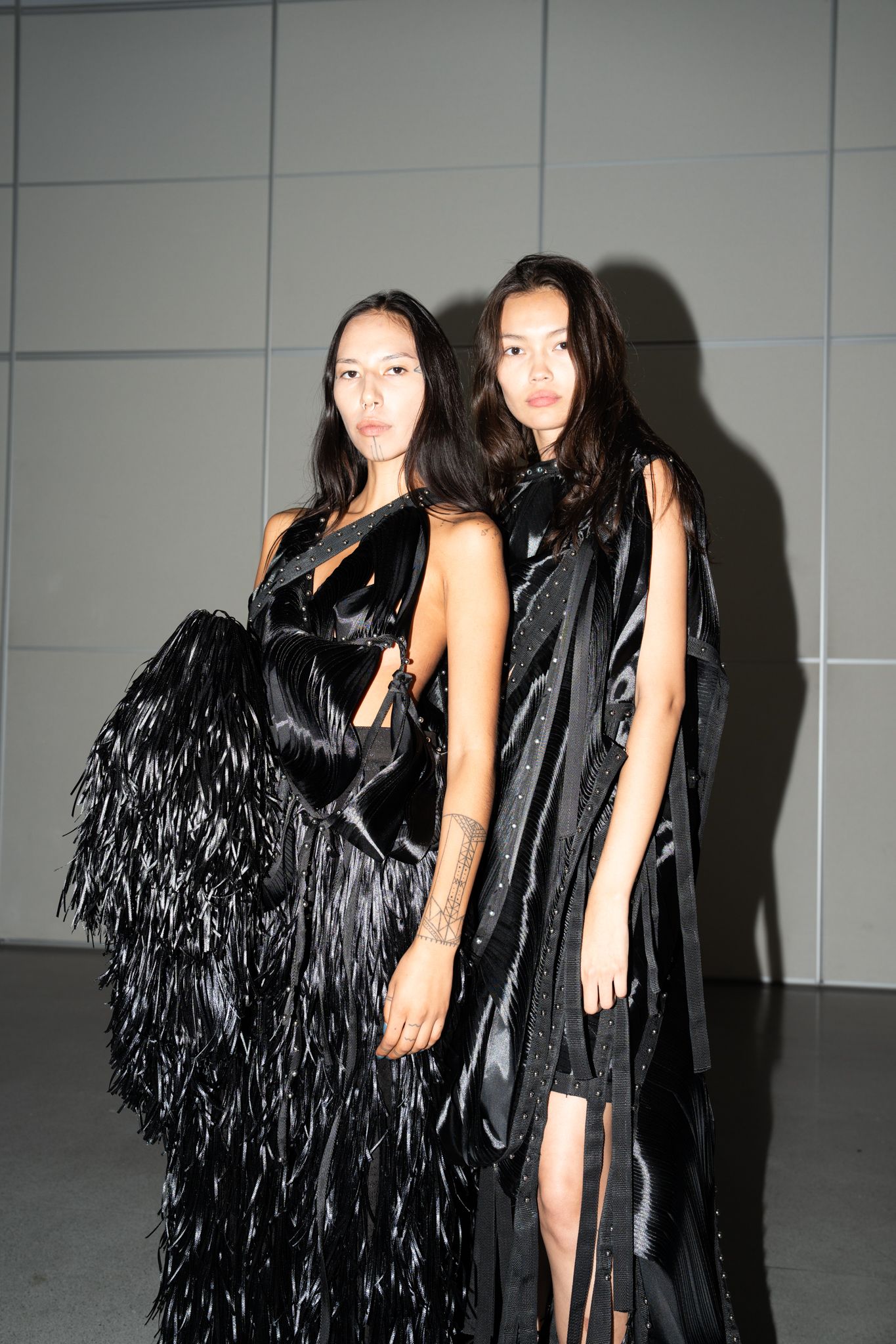 Two models post in black, draped garments featuring fringe, cutouts, and glossy textures against a neutral backdrop.
