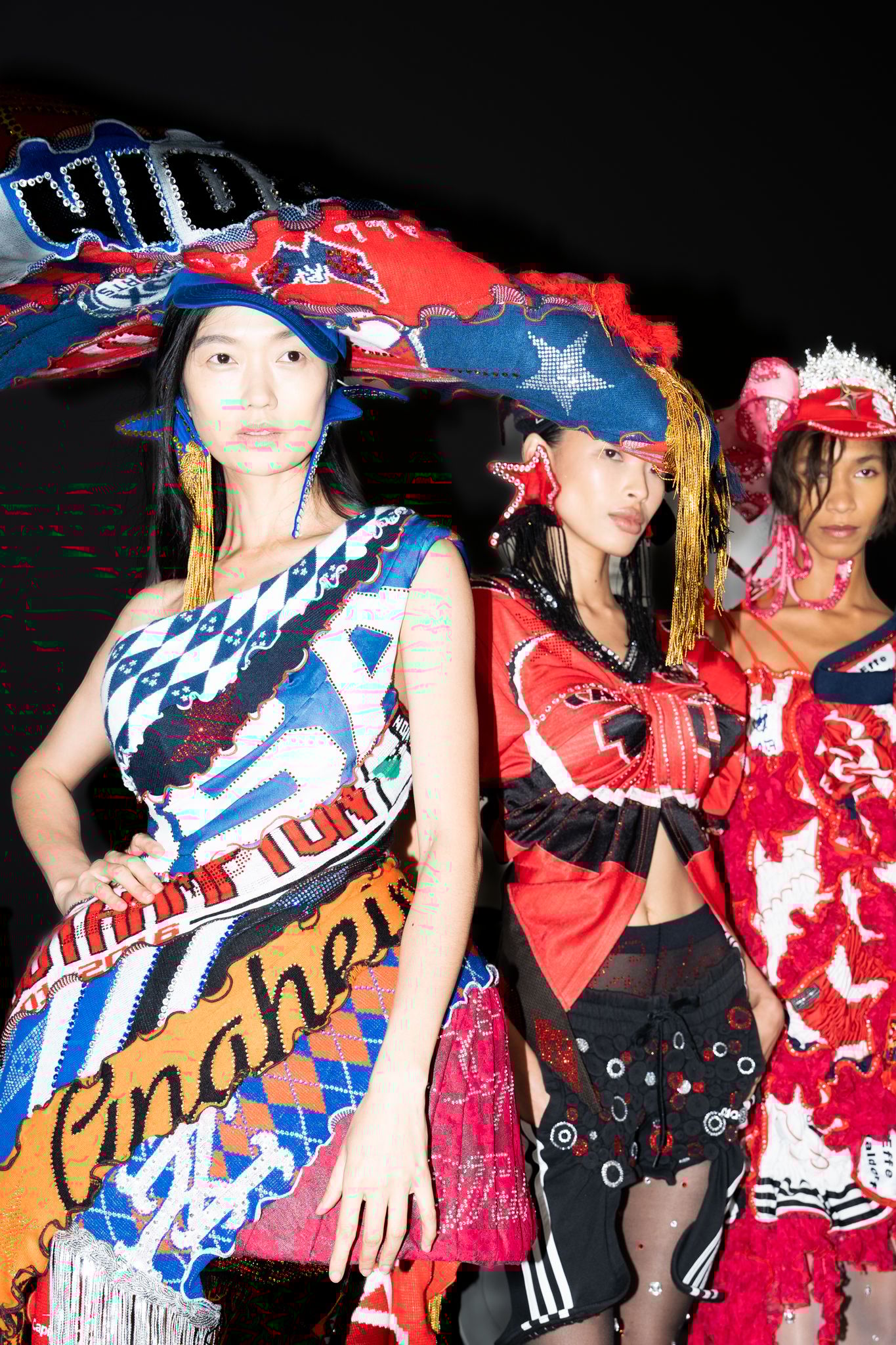 Three models wear bold red and blue garments with graphic patterns, layered embellishments and statement headpieces. 
