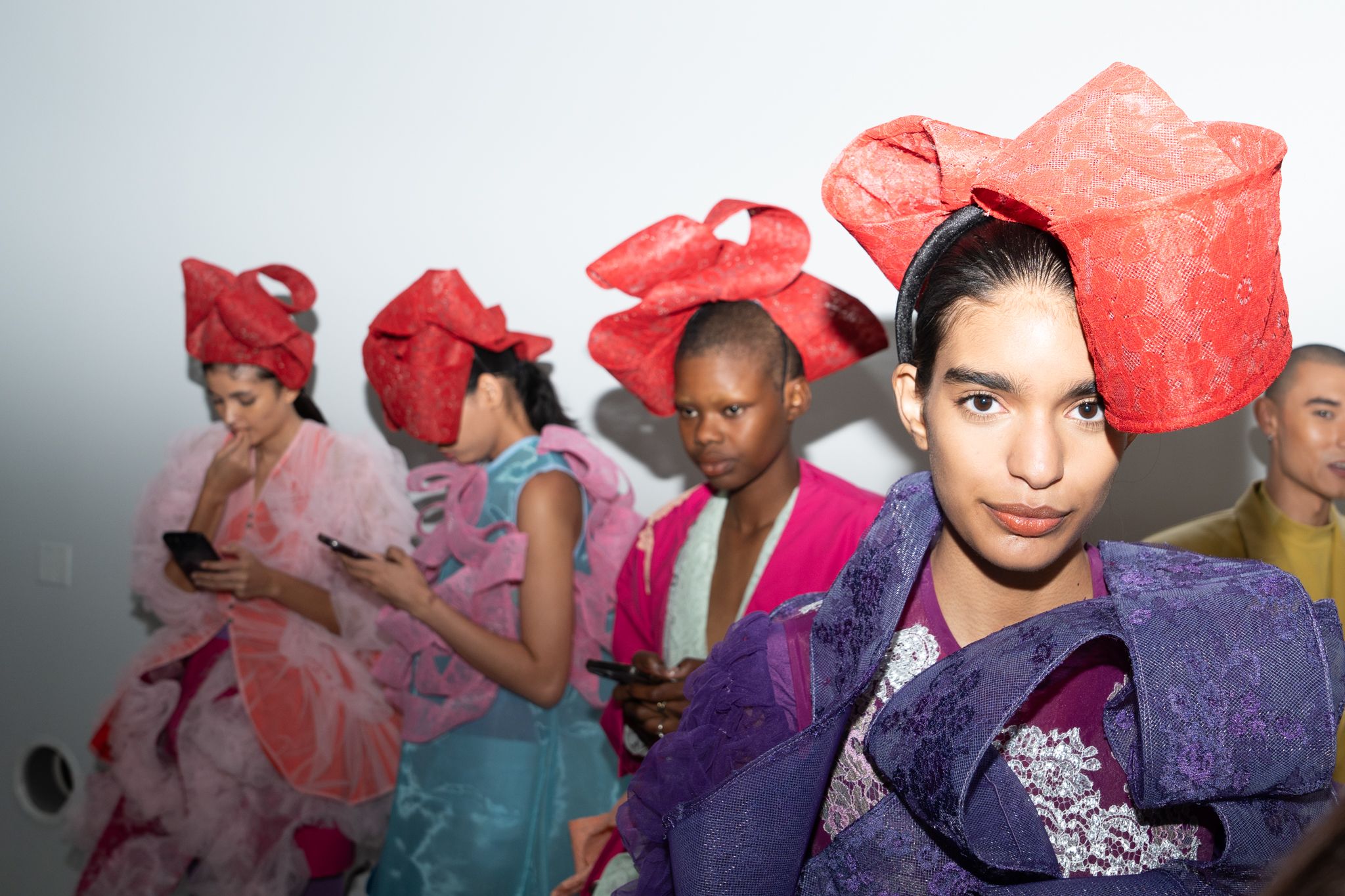 Models stand backstage wearing layered garments and oversized red headpieces, preparing before the runway.
