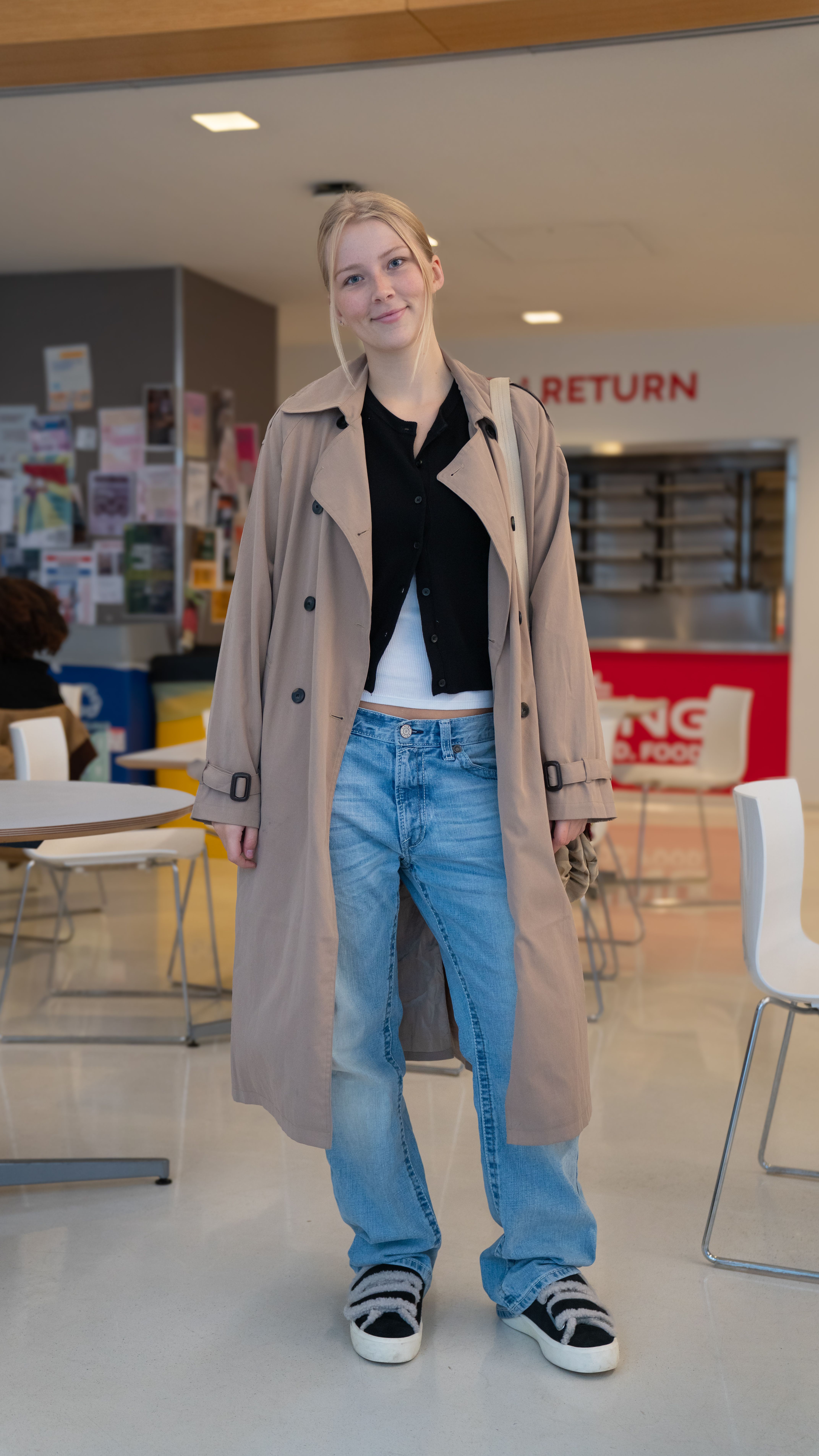 A girl standing indoors wearing a long beige trench coat over a black shirt, white top, light blue jeans, and black sneakers with white soles. They are smiling slightly in a casual setting with tables and chairs in the background.