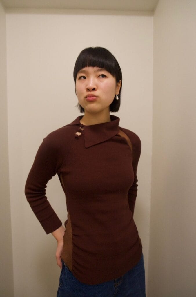 A girl with a black bob. She wears a maroon sweater with an asymmetrical, triangular collar that is buttoned up with knot-like buttons. She pouts her lips in a kissy-face expression.