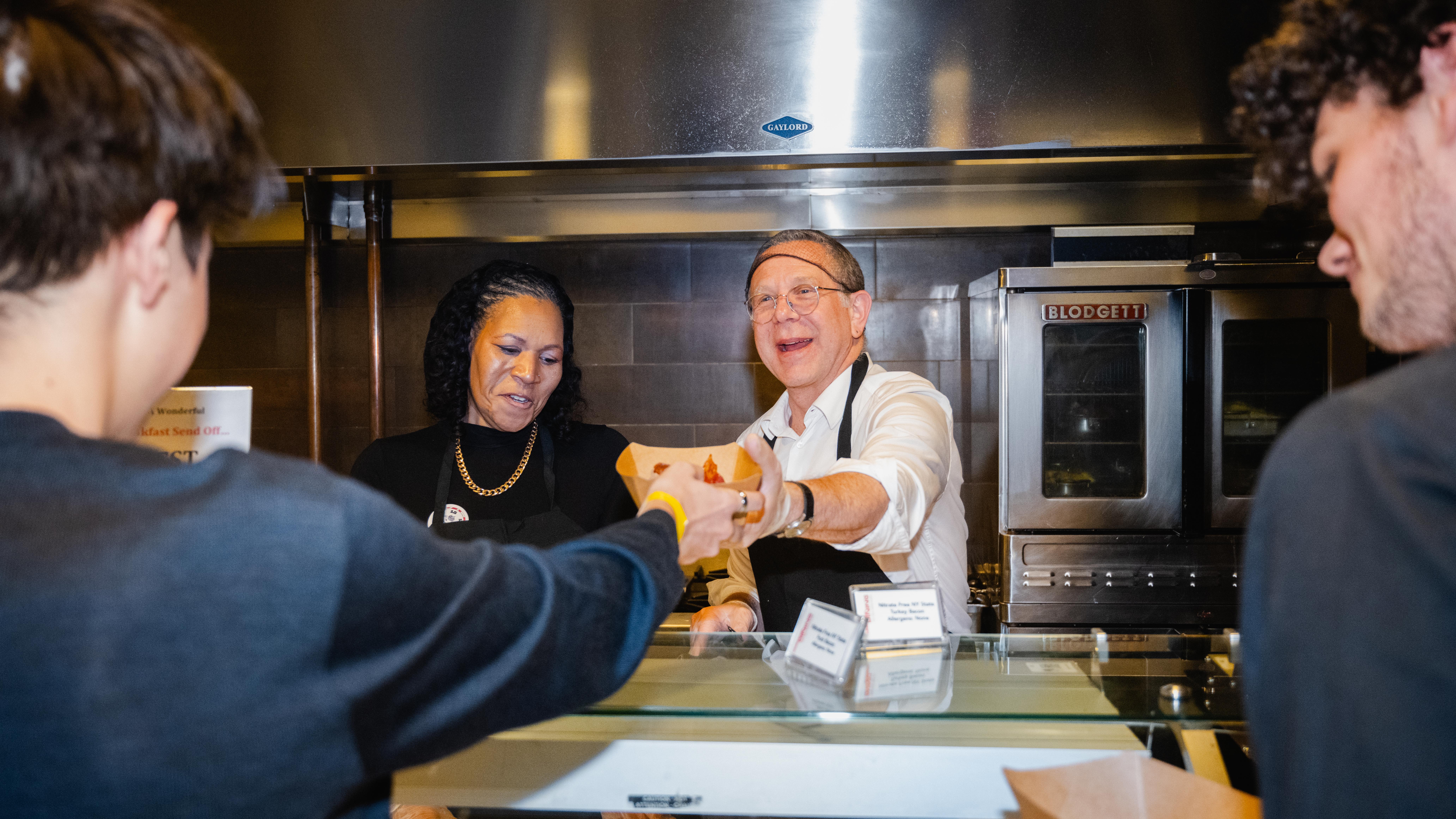 Man in a white shirt serving food to a man wearing a blue shirt. Next to the man serving the food is a woman in a black shirt.