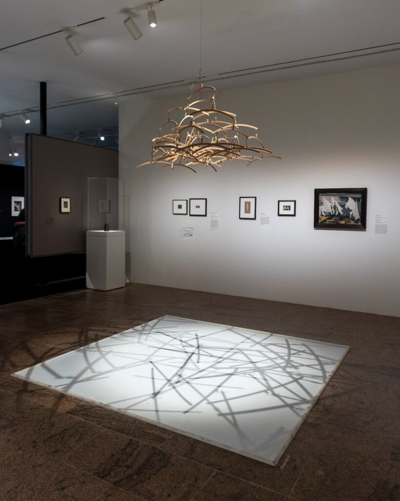 An image of a wooden sculpture in the center of the Man Ray exhibit, casting dark shadows on a white square on the ground.