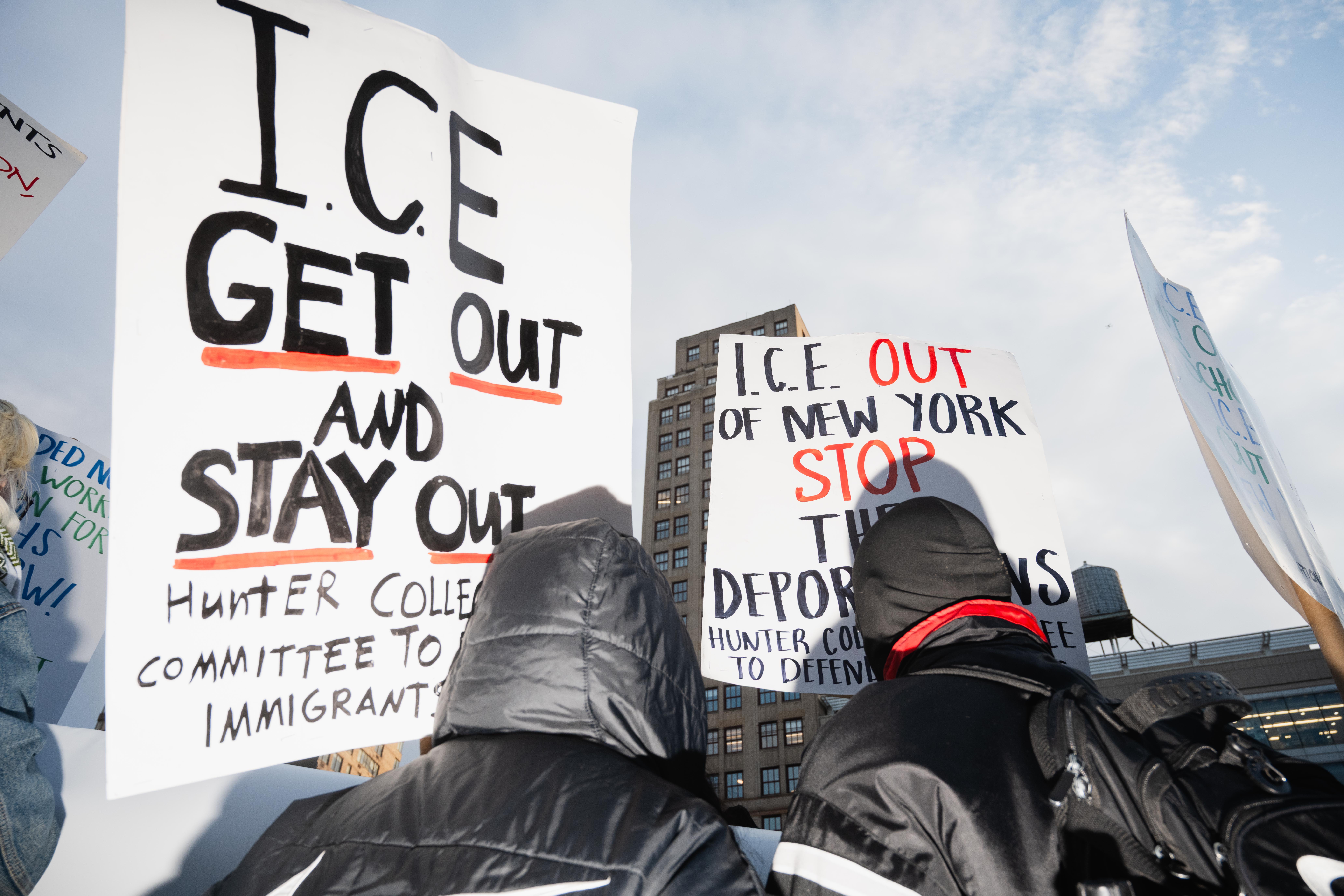 Two protesters hold large signs reading “ICE GET OUT AND STAY OUT” and “ICE OUT OF NEW YORK, STOP THE DEPORTATIONS.”