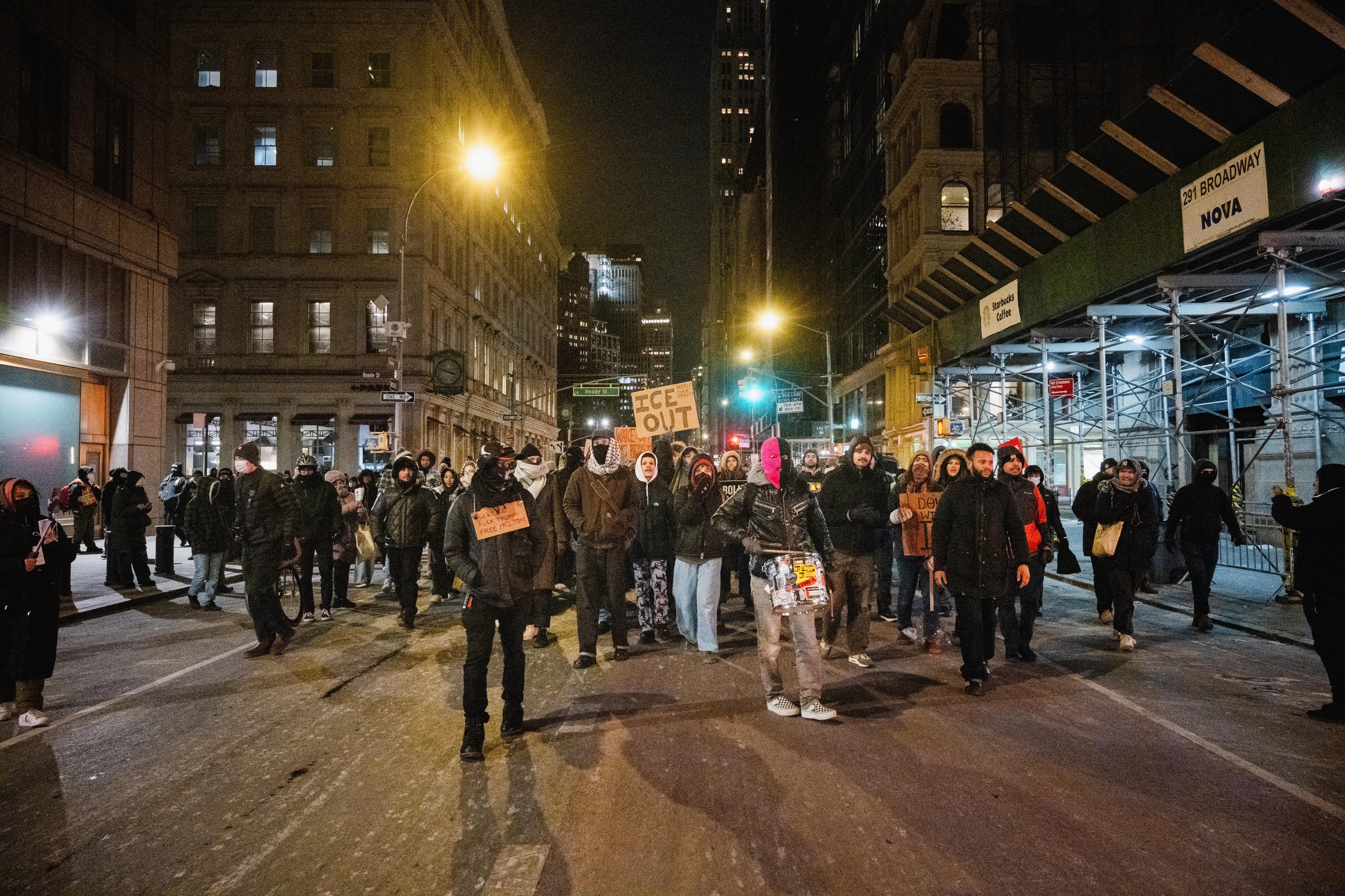A large group of protestors dressed in dark colors, with most of their faces covered, walk up a street at night.