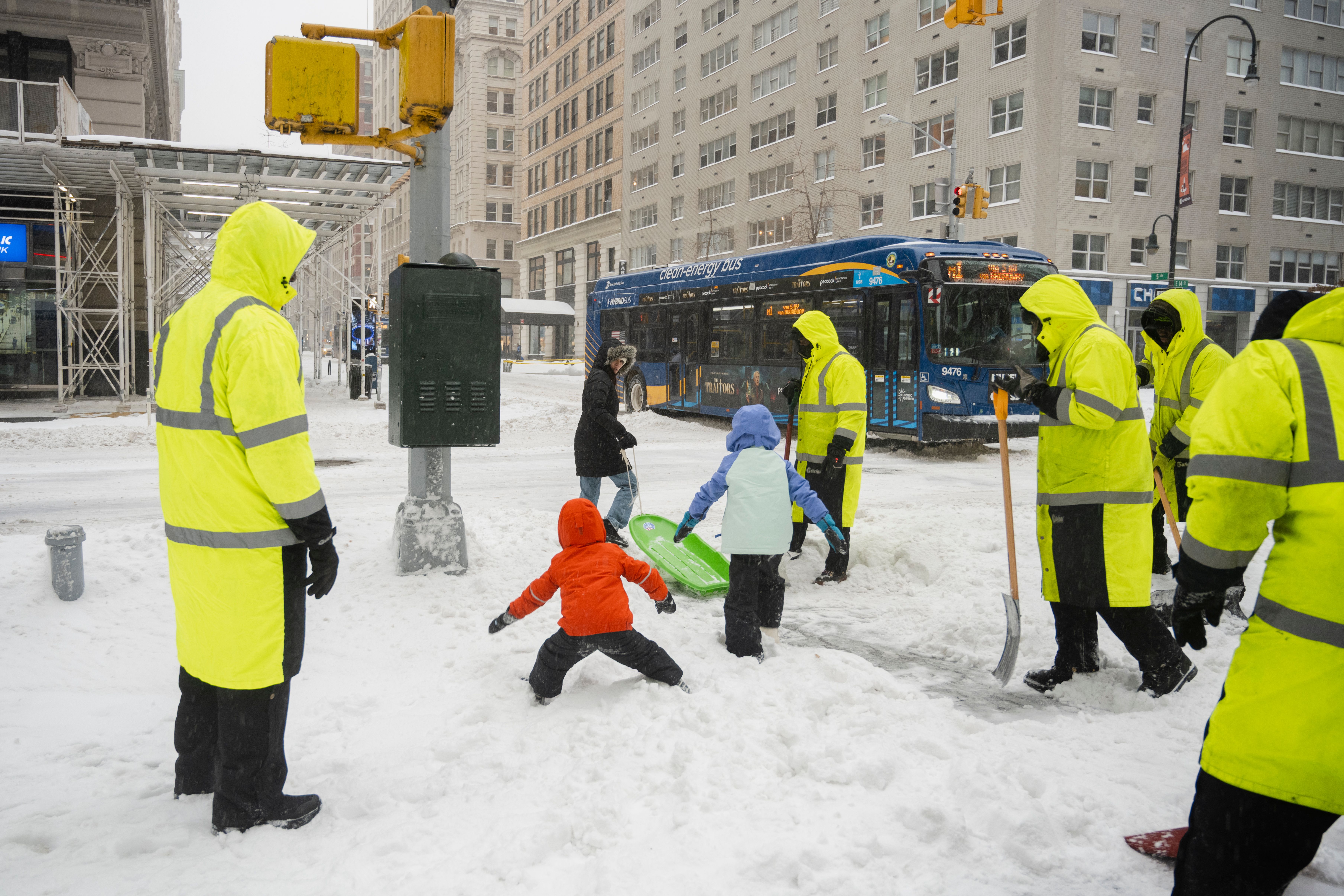 Children playing in a snowy street while people wearing high visibility suits stand nearby.