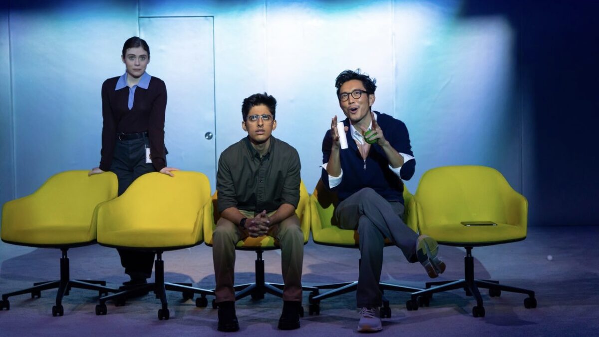 Alt text: Two men sitting on a row of five yellow office chairs, with a woman standing behind.