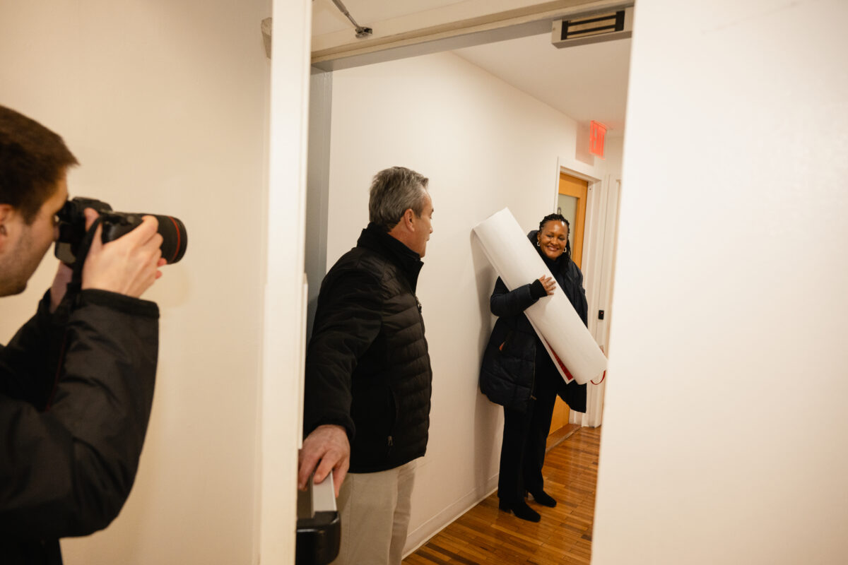 A woman is carrying a rolled up letter, while a man holds the door. A person takes a photo to the left.