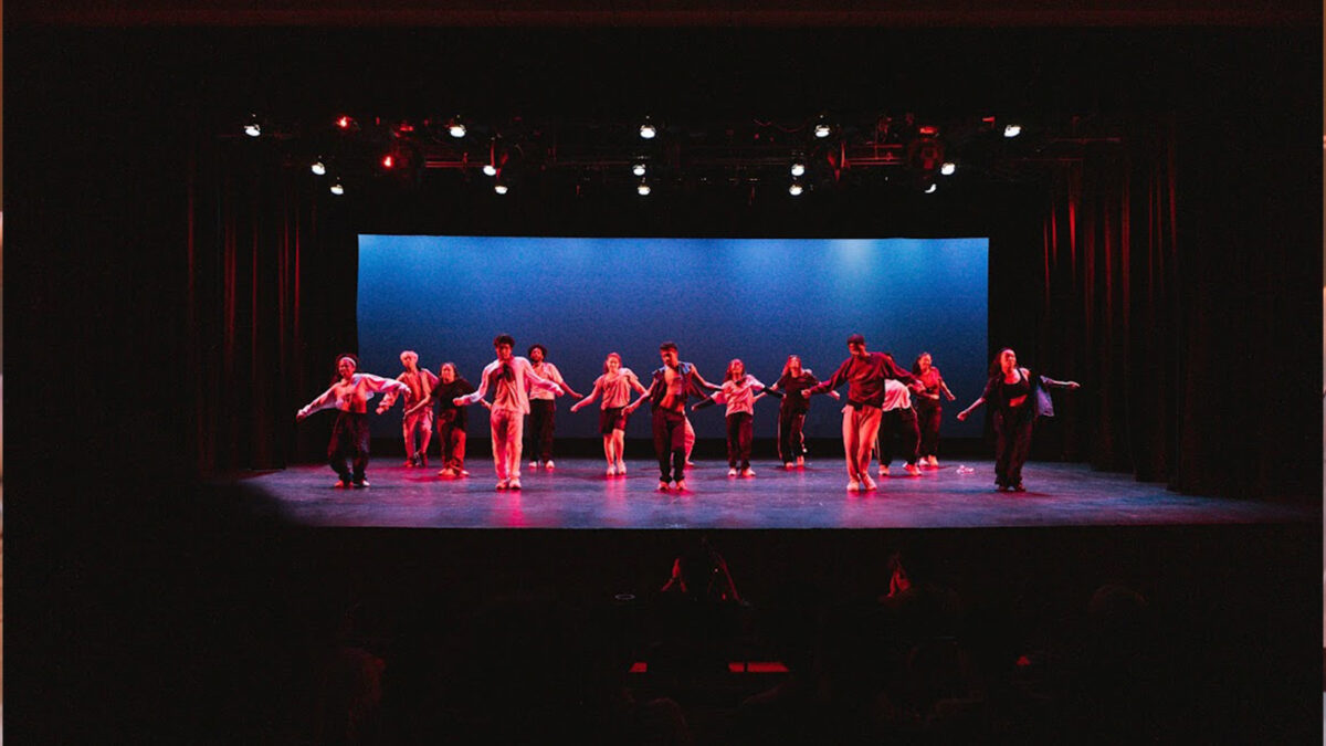 ALT TEXT: 14 dancers spread across a large dark stage.
