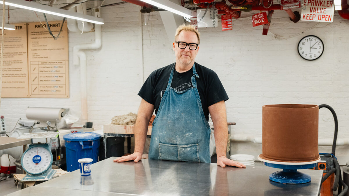 Andrew Cornell Robinson pictured with glasses stands at a metal desk wearing a denim smock and smiling at the camera.