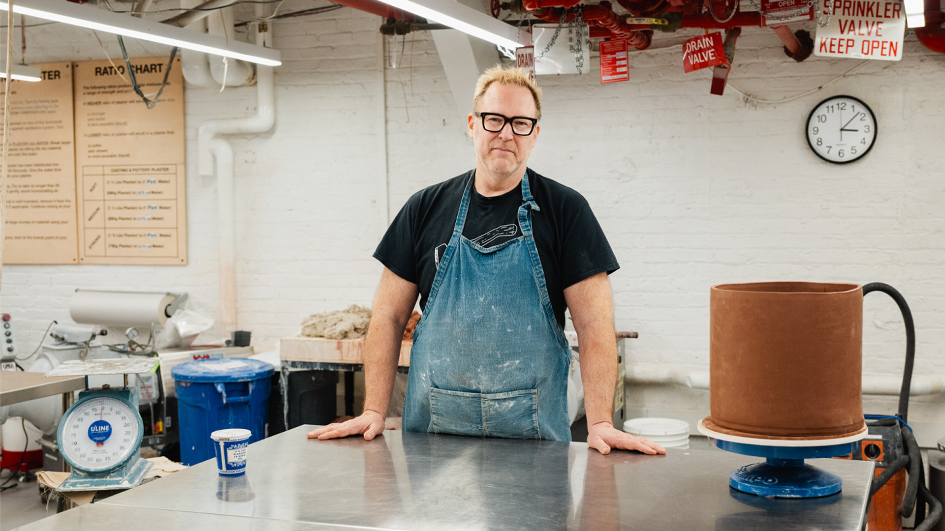 Andrew Cornell Robinson pictured with glasses stands at a metal desk wearing a denim smock and smiling at the camera.
