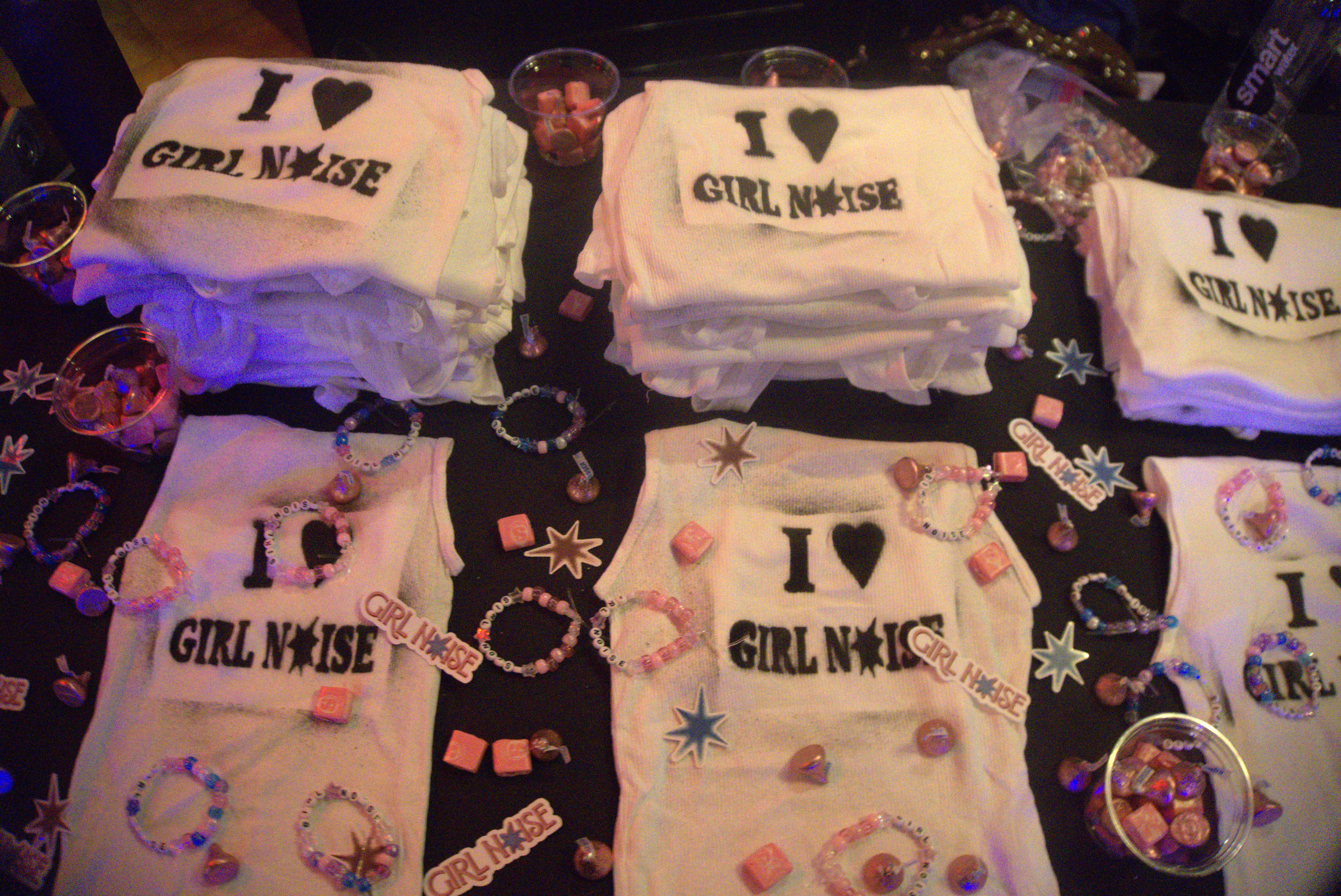 A photo of the Girl Noise merch table, which includes pink and purple friendship bracelets, tops with “I Heart Girl Noise” spray-painted on, and stickers.