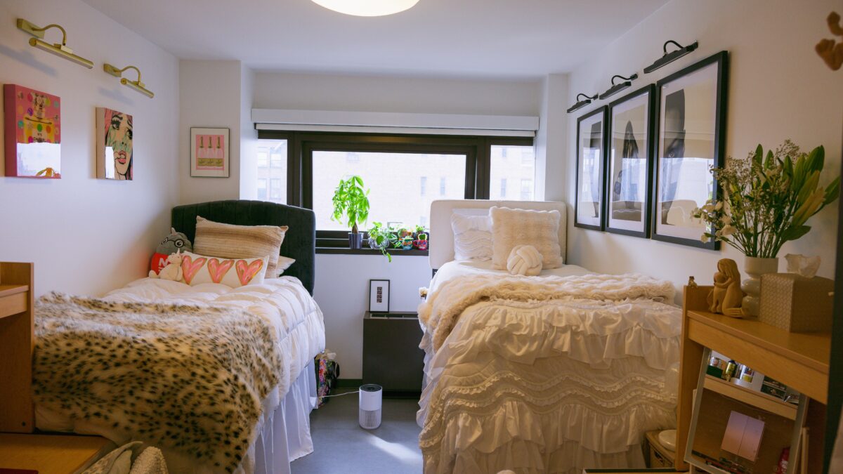 A fully furnished dorm room with two beds, two desks, and a wide window.