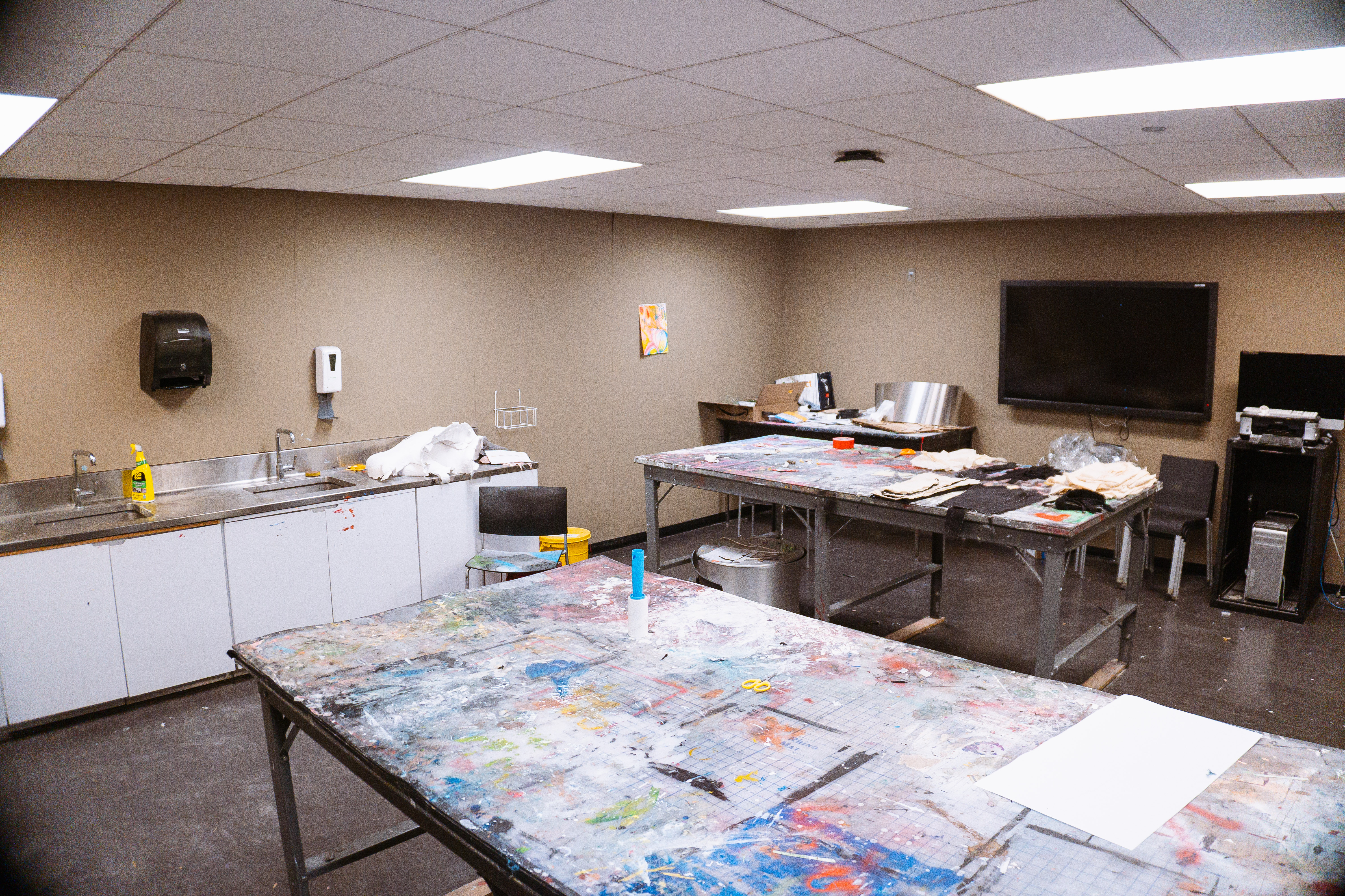 A room with paint-splattered tables, a countertop, a sink, art-supplies, and a large wall-mounted television. 
