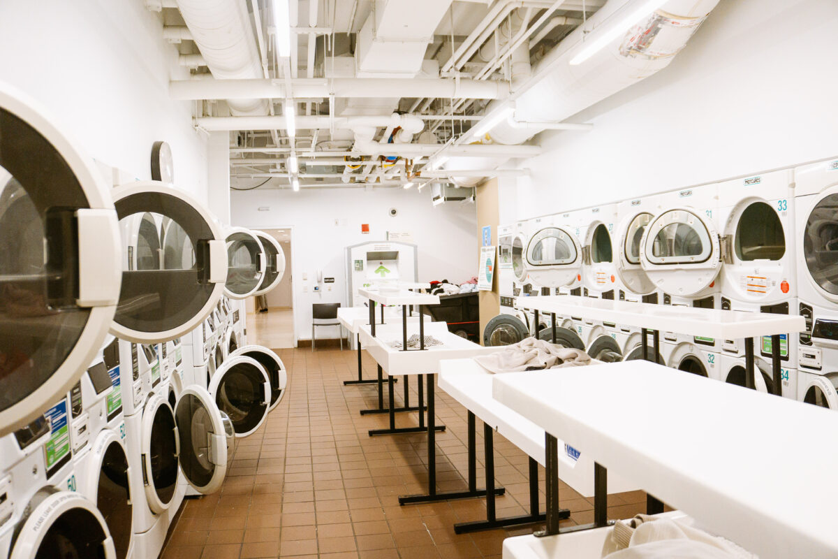 A long laundry room with tile floors. The walls are lined with stacked washer-dryer units and tables run down the center of the room.
