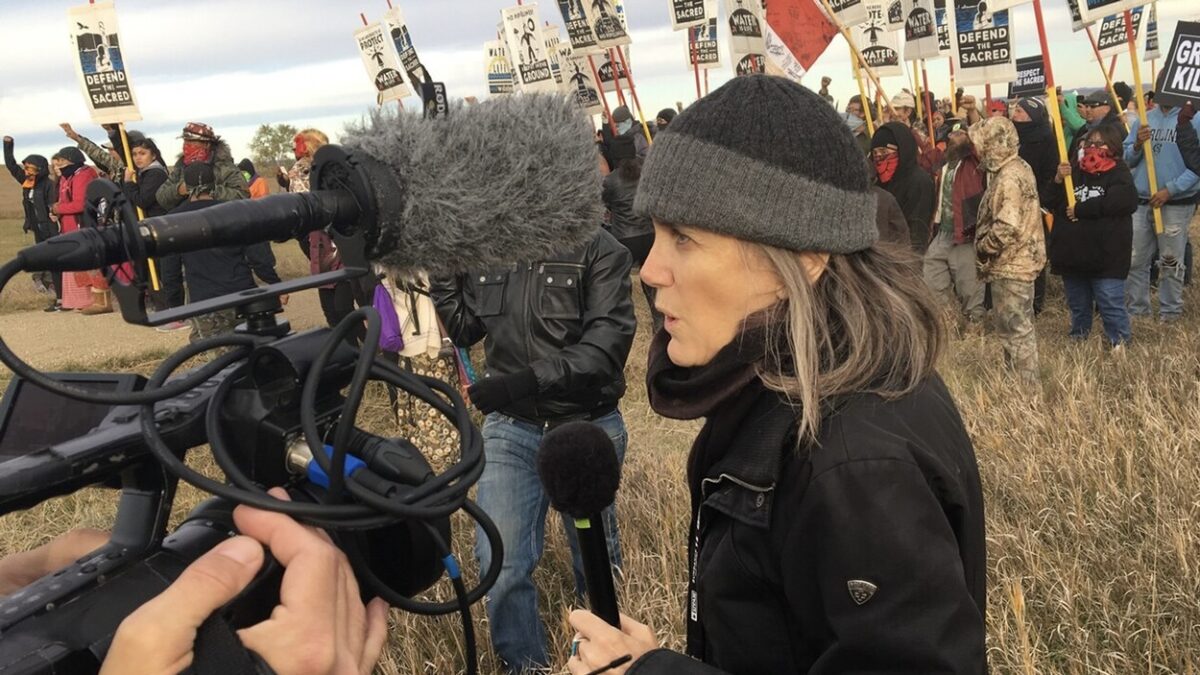 Amy Goodman stands in front of a line of protestors, speaking into a camera and mic.