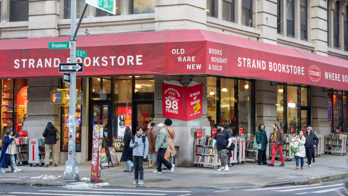 A street corner view of the Strand Bookstore with bookcases outside and front doors to the left. It is covered with a red awning that says “Strand Bookstore. Old, rare, new. Books bought & sold.”