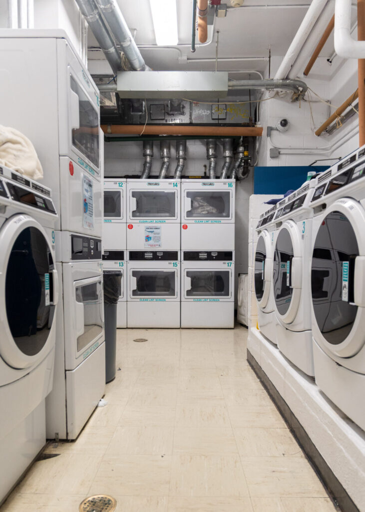 A basement laundry room with stacked washers and dryers, tile flooring, and fluorescent lighting.