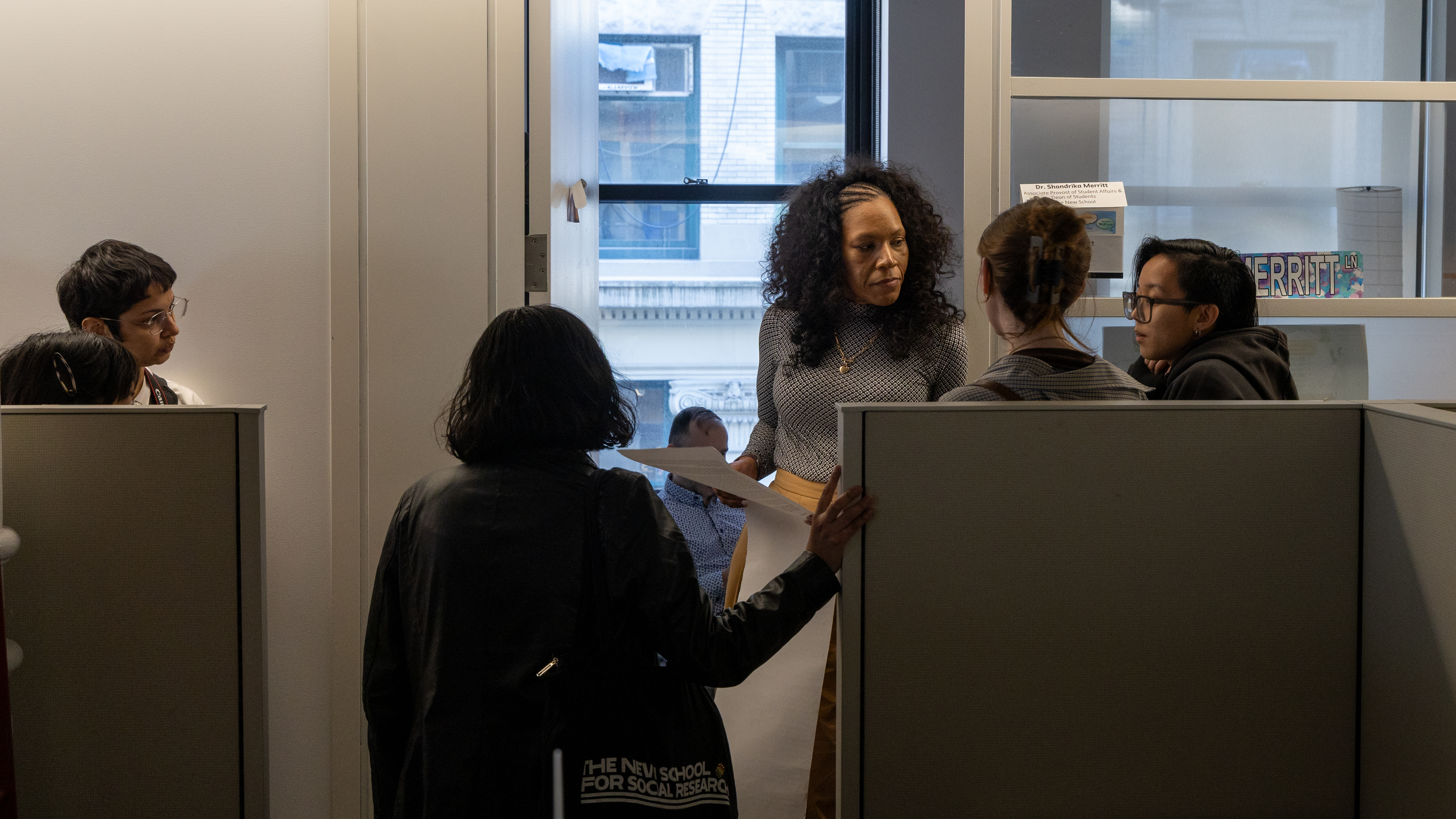 Multiple college students in an office, having a conversation with a woman who is holding paper.