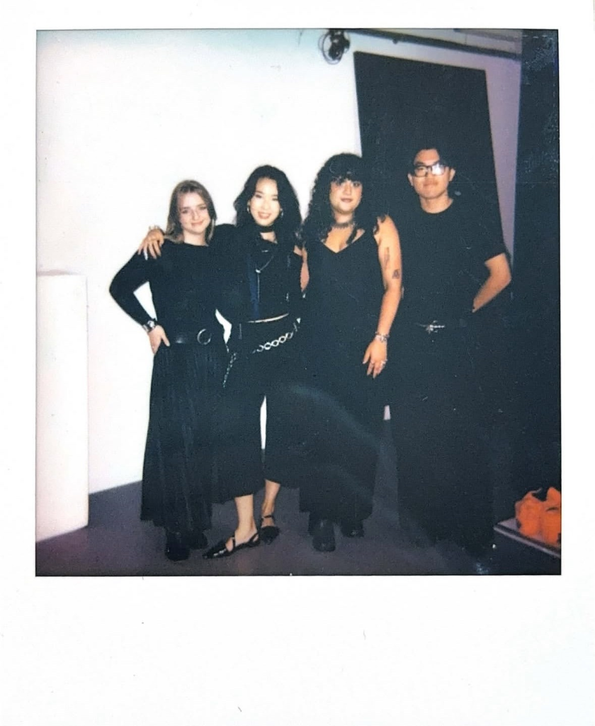 In a film style polaroid, four people dressed in black stand and pose for a photo.
